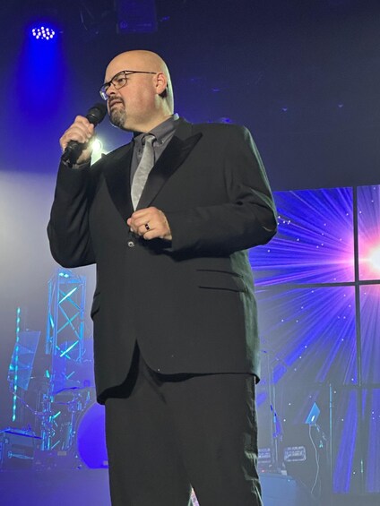 A man in a suit holding a microphone on a stage with a blue-lit background and a large screen displaying a structure. 2