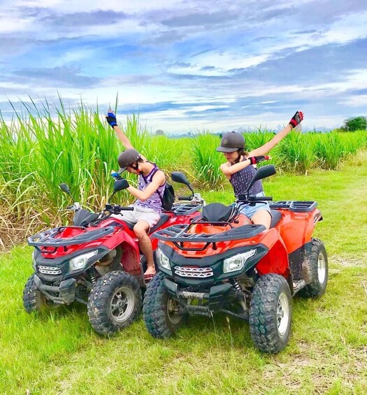 Two people on red all-terrain vehicles in a grassy field with tall crops in the background. 1