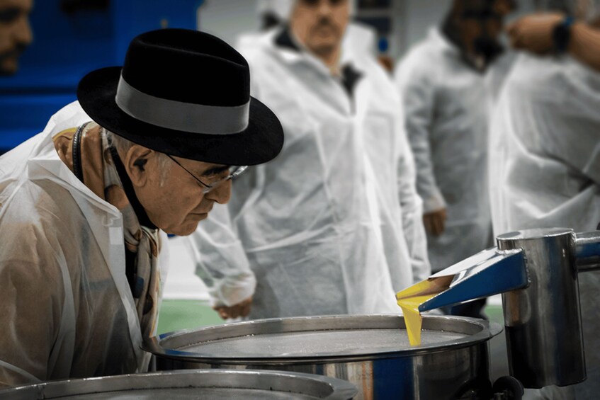 A person in a white lab coat and black hat working with a yellow substance in a stainless steel container. 3