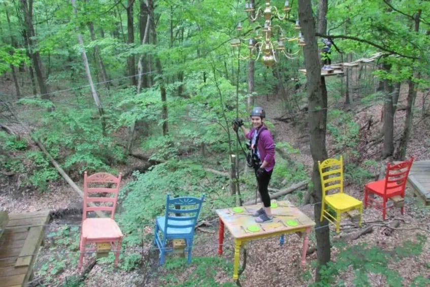 A person standing on a table in a forest with colorful chairs and a rope bridge. 5