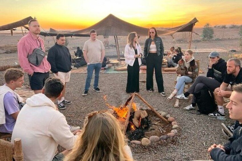 A group of people gathered around a campfire in a desert setting with tents in the background. 3