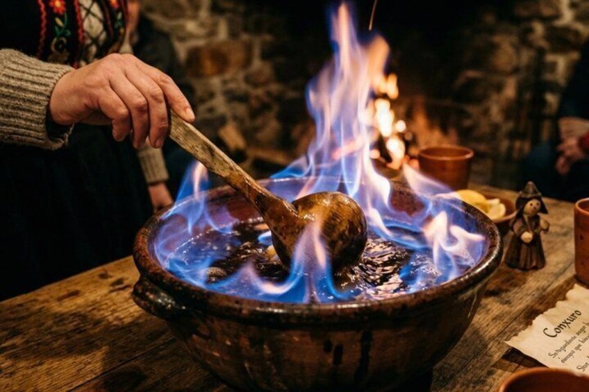 A person cooking with a large pot over an open flame, wooden spoon, and lit candles in the background. 1