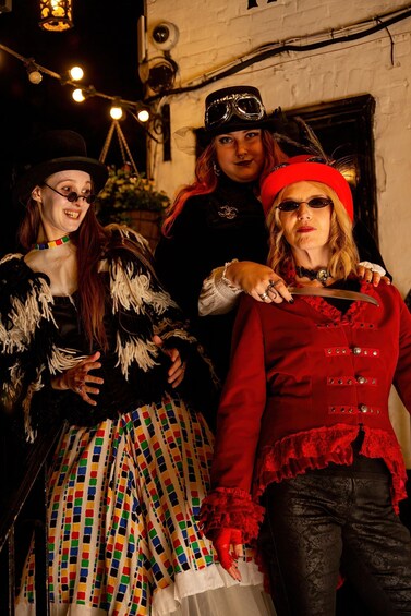 Three people in costumes, one holding a knife, in front of a building with a brick wall and string lights. 5