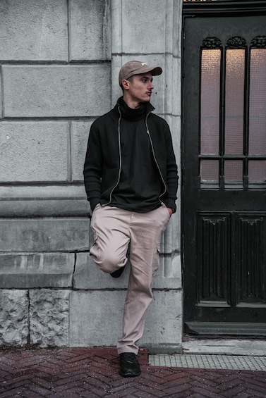 A person leaning against a stone wall, wearing a black jacket, beige pants, and a cap. 3