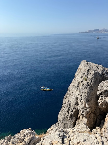 A kayaker in a blue kayak on a calm sea, with rocky cliffs in the foreground and a distant coastline. 4