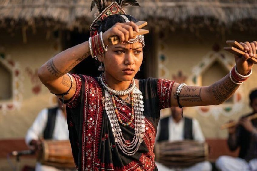A person in traditional attire with elaborate jewelry and a headdress, standing outdoors with a thatched-roof building in the background. 1