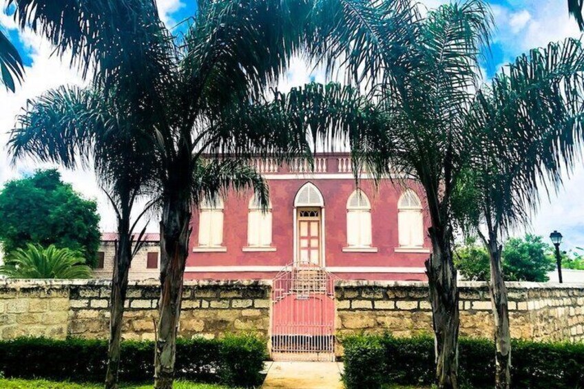 A red building with arched windows and a central door, surrounded by palm trees and a stone wall. 2