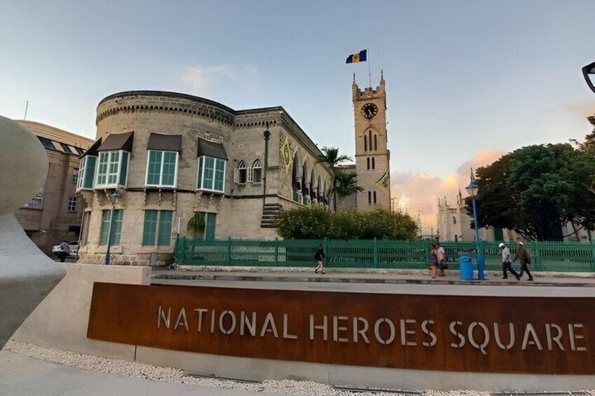 A historic building with a clock tower, labeled 'NATIONAL HEROES SQUARE,' and a sign in front. 5