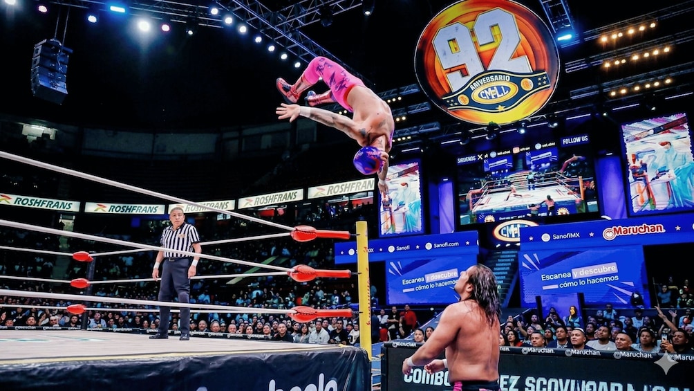 A wrestling match with a referee, a wrestler performing a flip, and a large '92' anniversary sign. 1
