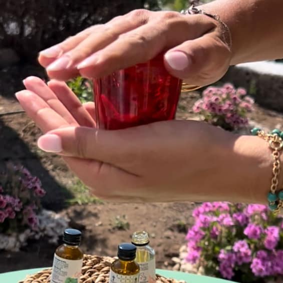 Hands holding a red glass, with small bottles and flowers in the background. 1
