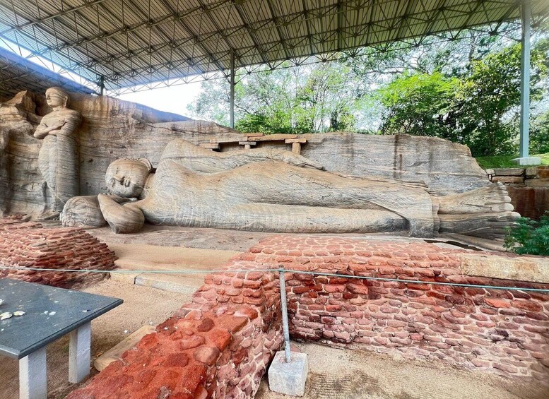 A large stone Buddha statue under a metal roof, surrounded by brick structures and greenery. 2