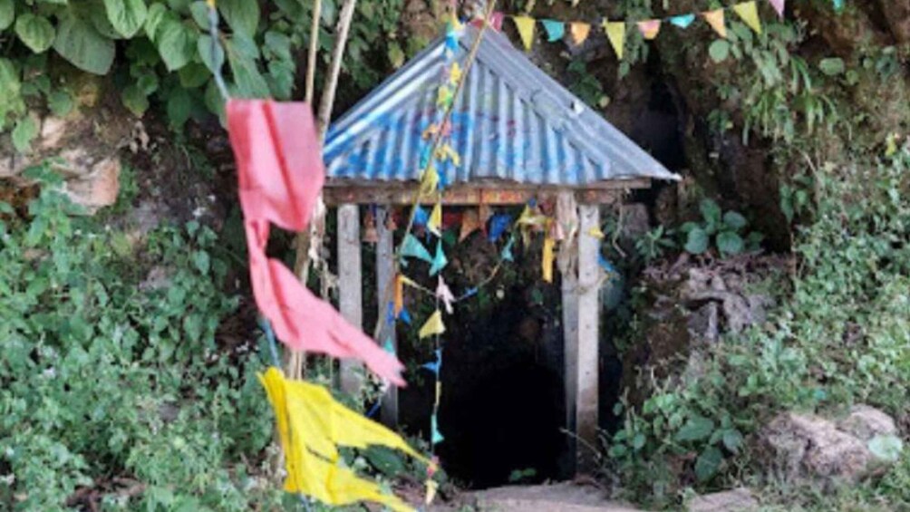 A small shrine with a corrugated metal roof, surrounded by greenery and adorned with colorful flags. 3