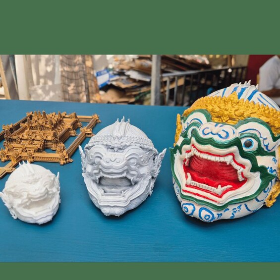 A collection of cultural artifacts including a detailed wooden model, a white 3D-printed mask, and a colorful painted mask. 2