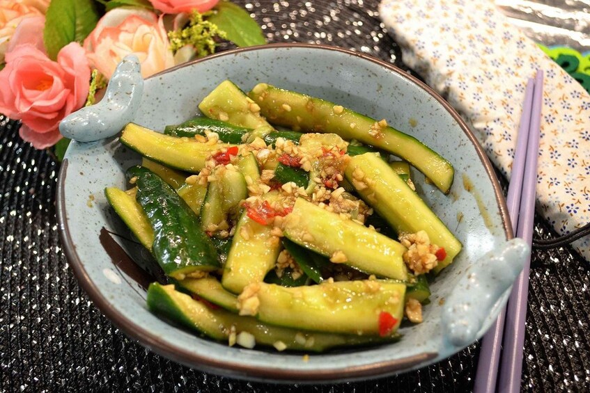A bowl of sliced green vegetables with nuts and red peppers, accompanied by chopsticks and a decorative ceramic piece. 1