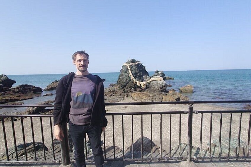A person standing on a rocky beach with a railing, a large rock formation, and the sea in the background. 2