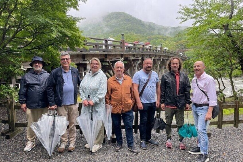 A group of people standing in front of a wooden bridge with a river and mountains in the background. 4