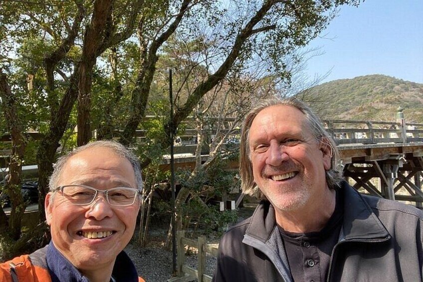 Two people standing outdoors with a wooden bridge and trees in the background. 3
