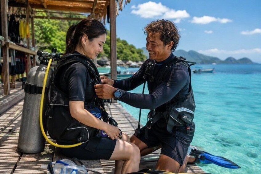 Two people in scuba diving gear on a dock with clear blue water and a tropical landscape in the background. 2