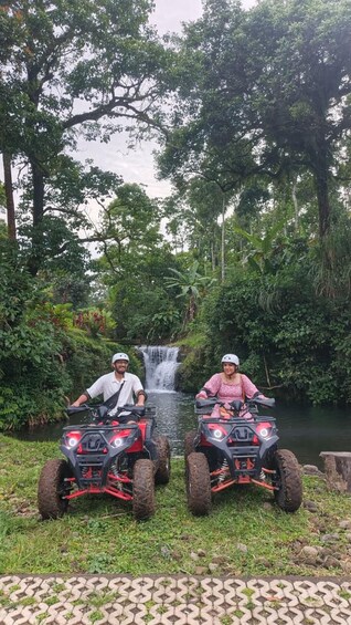 Twee personen op quadbikes in een weelderig, groen gebied met een waterval op de achtergrond. 3