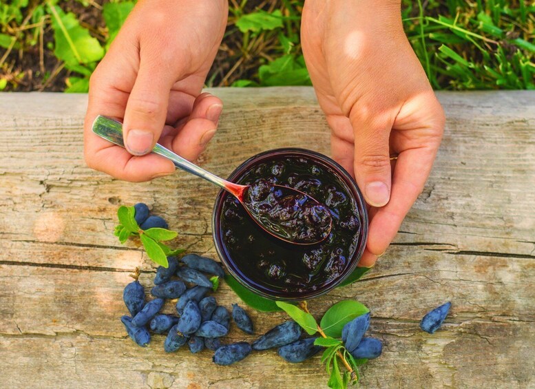 A person holding a bowl of blueberry jam with a spoon, surrounded by blueberries and leaves on a wooden surface. 5
