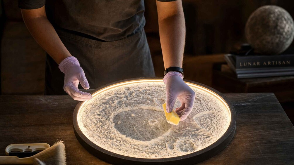A person wearing gloves, working on a circular, illuminated surface with a powdery substance. 4