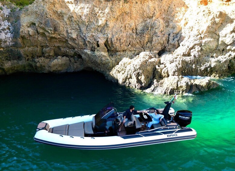 A white inflatable boat with 'BRIG' branding, two people, and a Suzuki outboard motor, anchored near a rocky coastline. 3