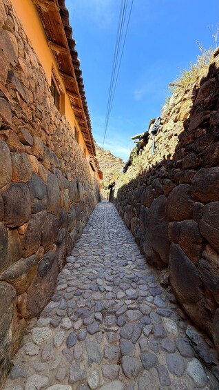 A narrow cobblestone street flanked by stone walls and a building with a wooden roof. 2