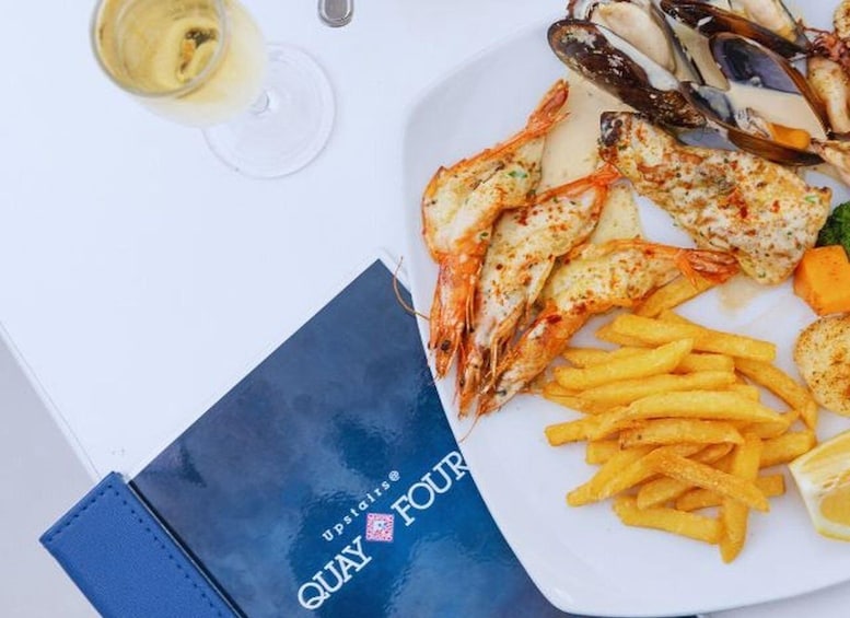 A plate of seafood including shrimp and mussels, accompanied by French fries and a glass of white wine, with a blue napkin featuring 'QUAY FOUR' logo. 2