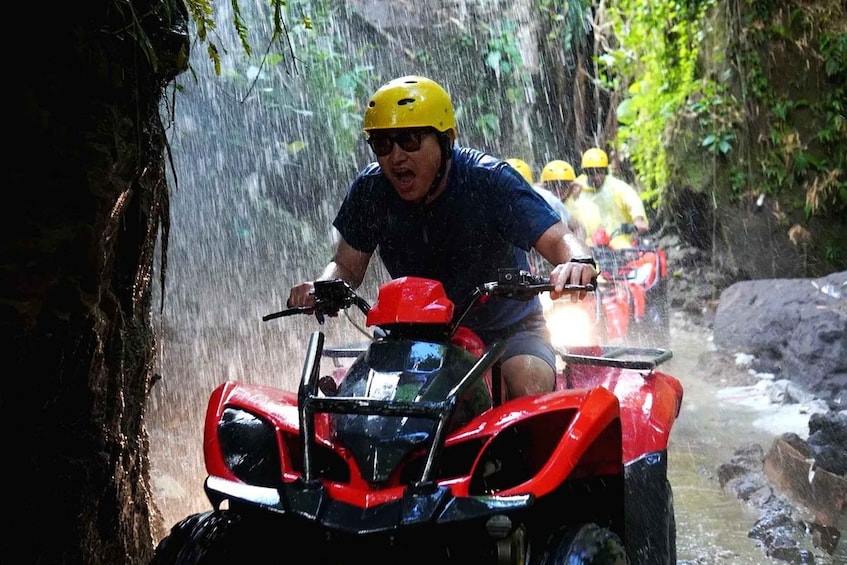A person riding a red ATV through a narrow, rocky water channel surrounded by greenery. 4