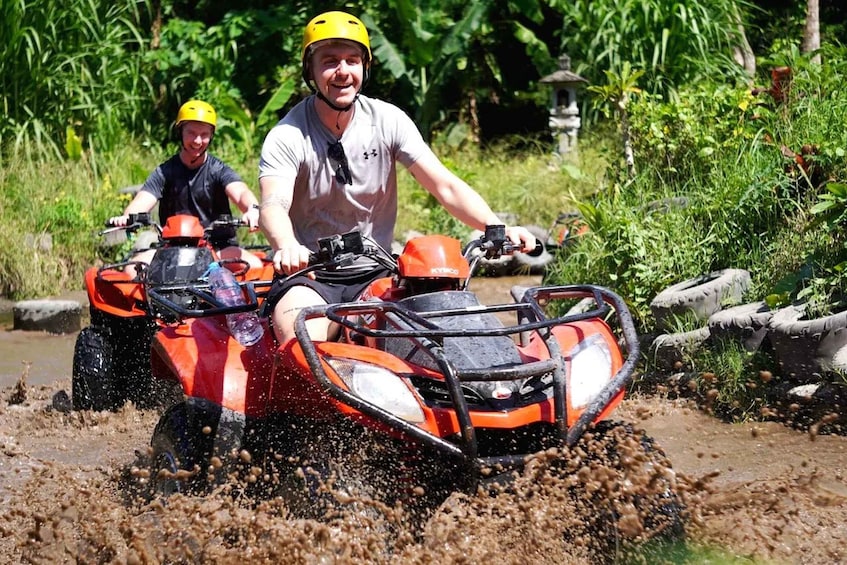 Two people riding an ATV through a muddy area with vegetation in the background. 2