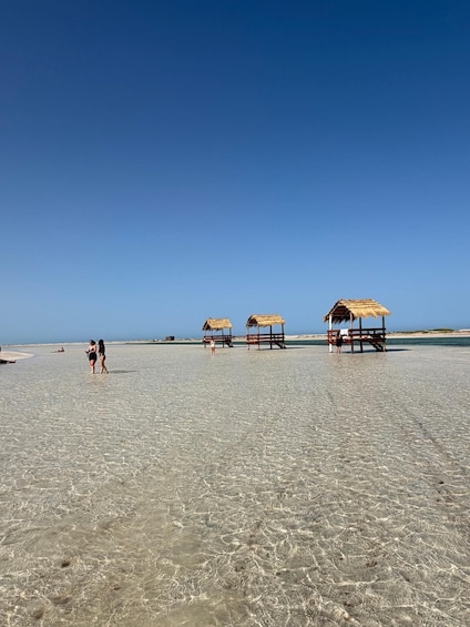 A beach with clear shallow water and three thatched-roof structures. 4