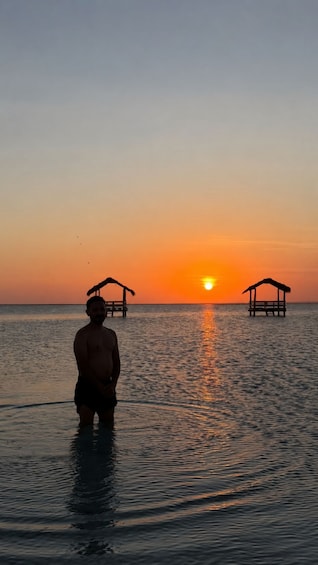 A person standing in calm water at sunset with two thatched-roof structures on the horizon. 3