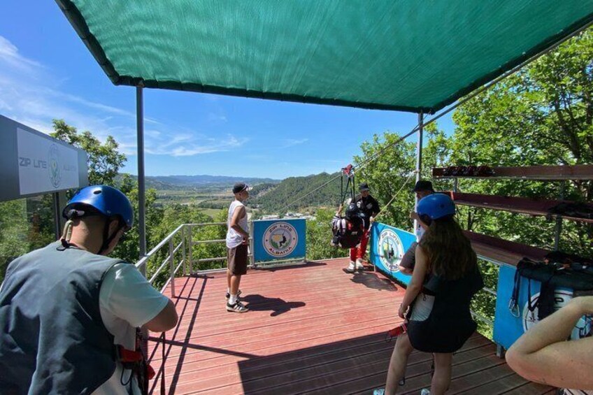 A group of people on a wooden platform with a scenic view, wearing helmets, and a sign with a circular emblem. 3