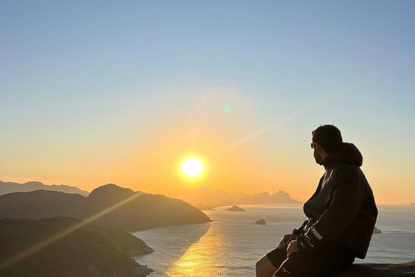 A person sitting on a rock overlooking a body of water with mountains in the background during sunset. 1