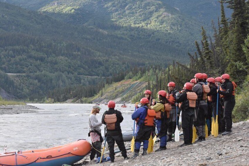 A group of people wearing helmets and life jackets, preparing for a water activity on a riverbank. 4