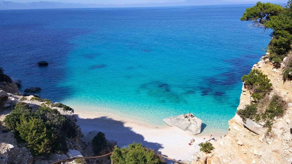 A coastal landscape with clear turquoise waters, a sandy beach, rocky outcrops, and sparse vegetation. 3