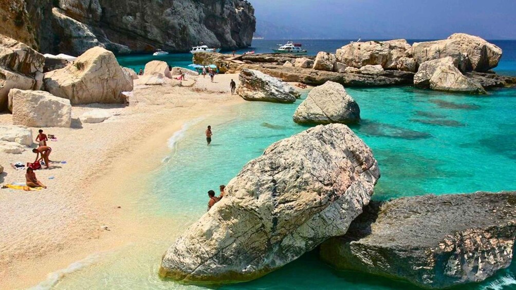 A rocky beach with clear turquoise water, large boulders, and a few people sunbathing. 2