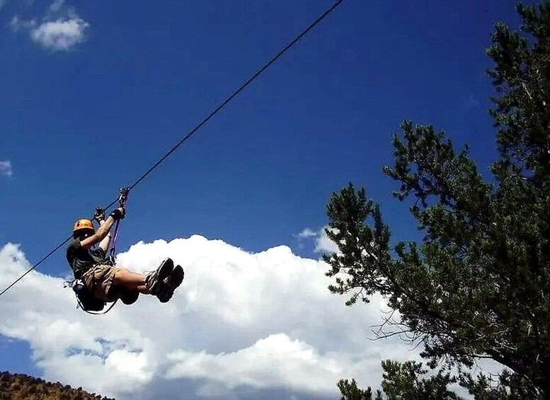 A person ziplining through a clear blue sky with clouds, trees, and a mountain in the background. 2