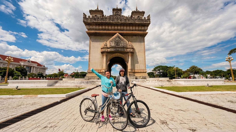 Two people on bicycles in front of a large, ornate gateway with a clear sky above. 1
