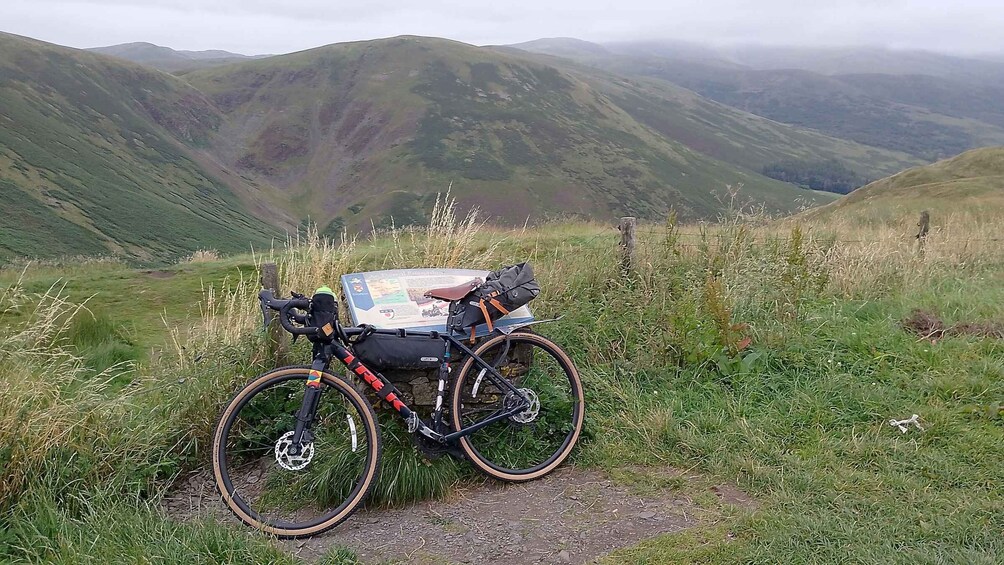 A mountain bike with a map and gear on the back rack, parked on a grassy path with hills in the background. 1