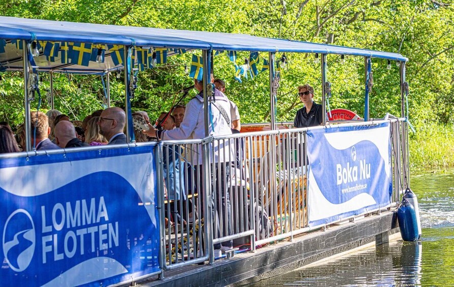 A boat with 'LOMMA FLOTTEN' and 'Boka Nu' branding, passengers, and a water body with greenery. 2