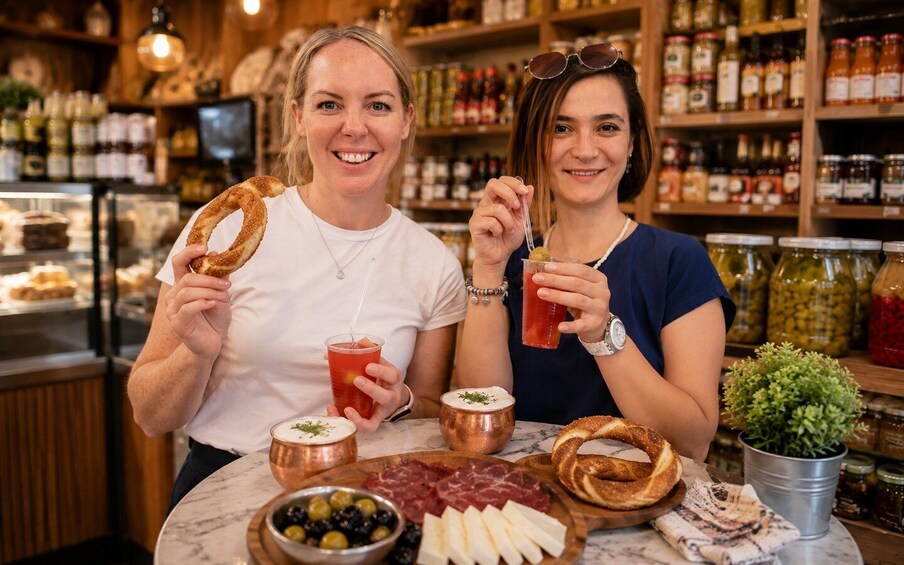 Two people holding pretzels and drinks, with a table of food and drinks in a restaurant. 2