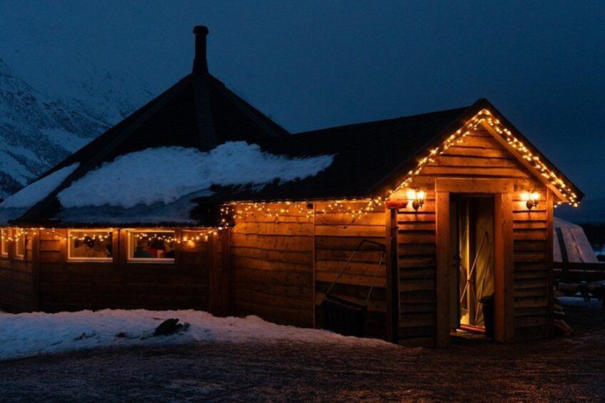 A wooden cabin with snow on the roof, surrounded by snow, and lit by string lights. 2