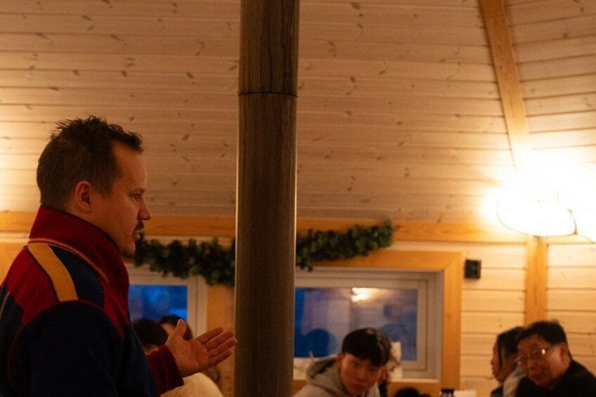 A wooden interior with a person standing and gesturing, a window, and a wall-mounted light fixture. 4