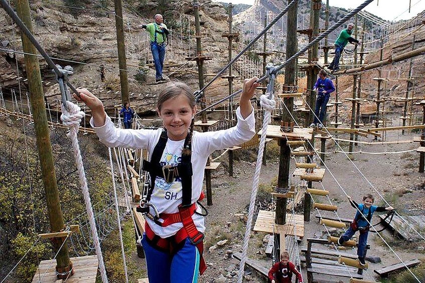 A person on a rope bridge with safety harnesses, surrounded by wooden structures and rocky terrain. 4