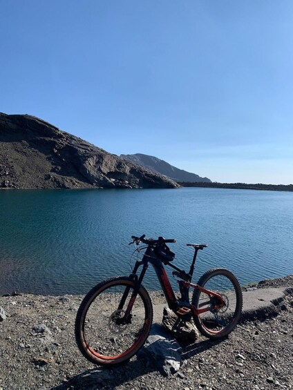 A mountain bike parked on a rocky shore by a calm lake with mountains in the background. 1