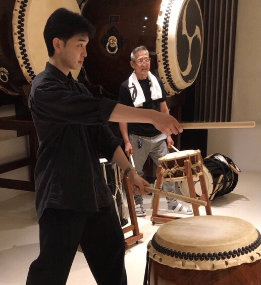 A person playing a drum, with a large drum set in the background. 2