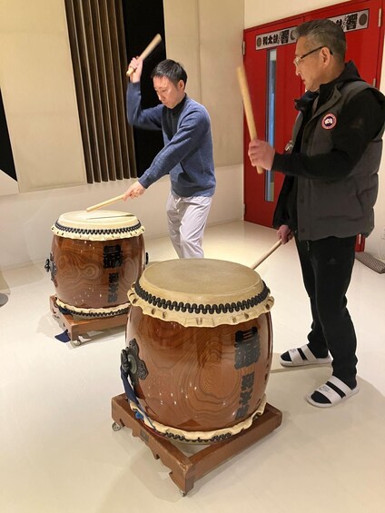 Two individuals playing drums in a room with a red door and a black curtain. 1