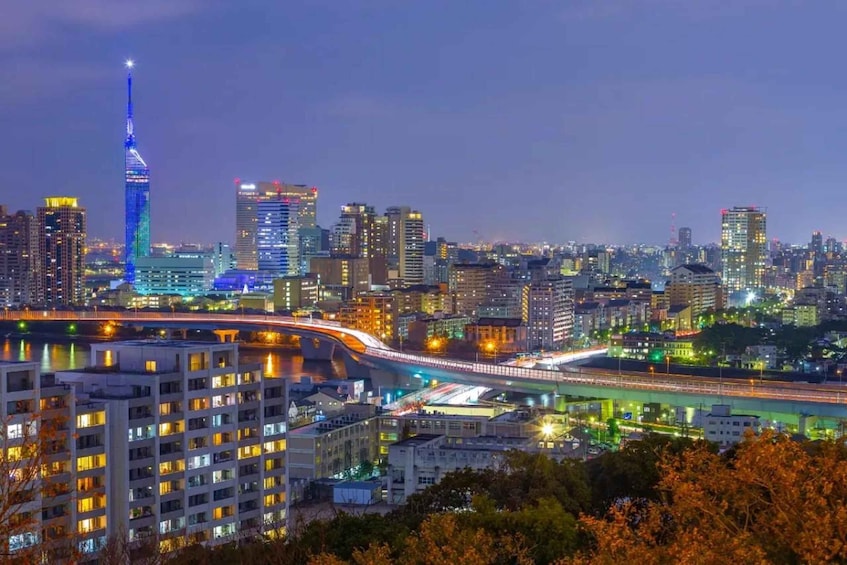A cityscape at dusk with illuminated buildings, a prominent tower, and a bridge. 4