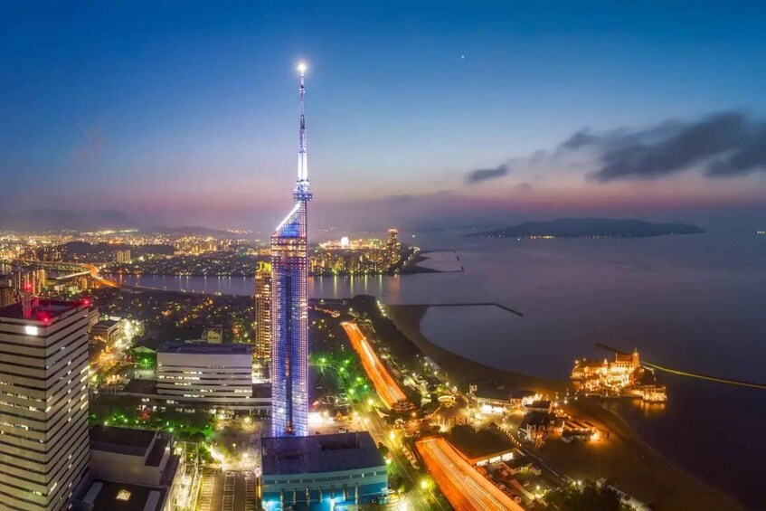 A cityscape at dusk with a prominent illuminated tower, a waterfront, and surrounding buildings. 1